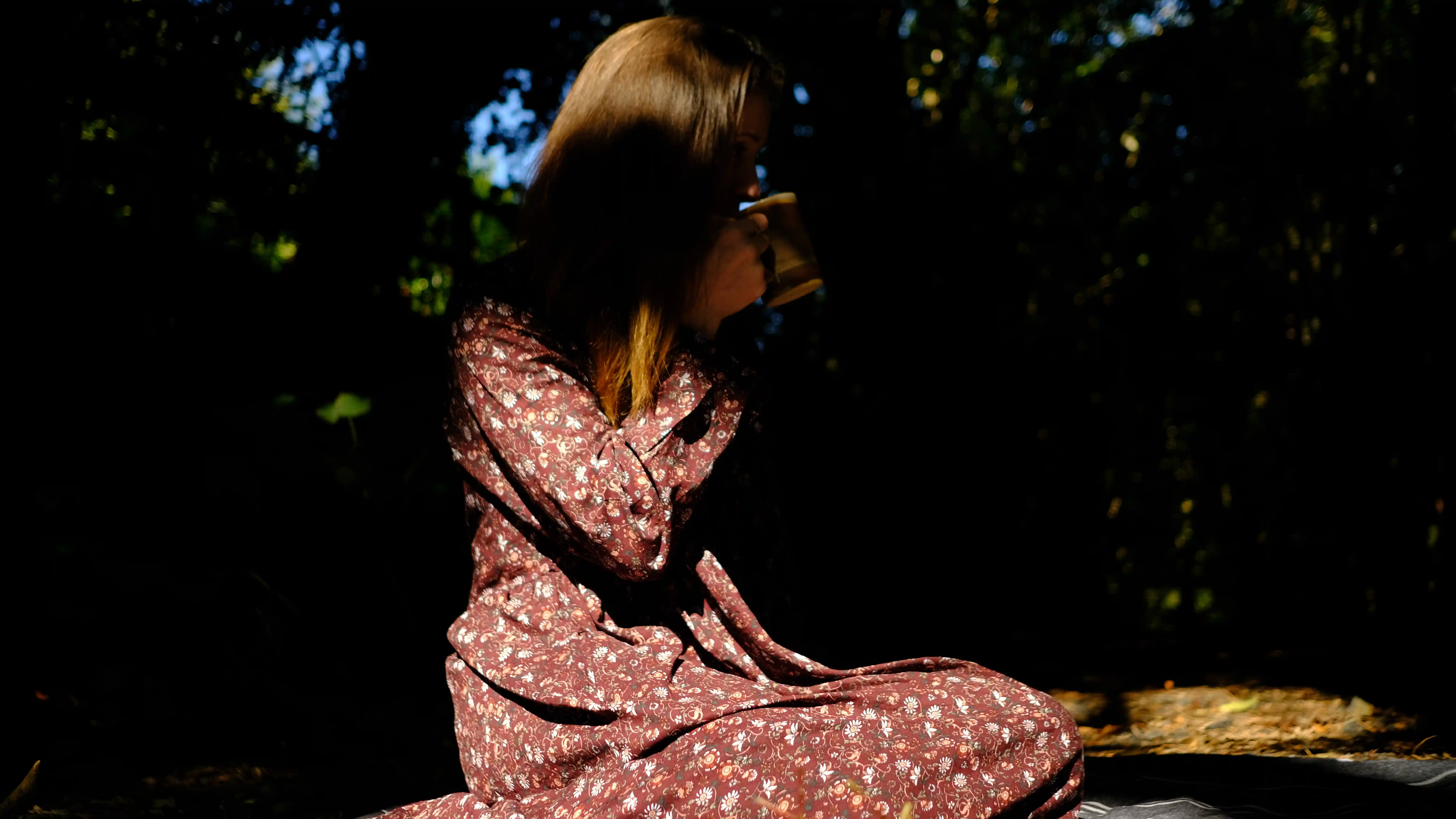 A woman sitting in the shadows of trees in nature drinking tea